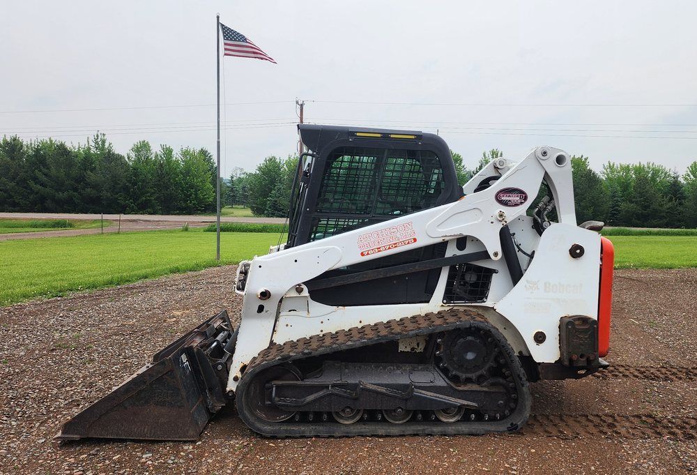 White and black track skid steer with a bucket on gravel, American flag in background.