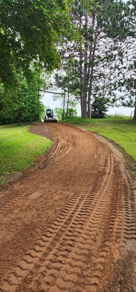 A muddy road winds towards a small bulldozer near trees and water.