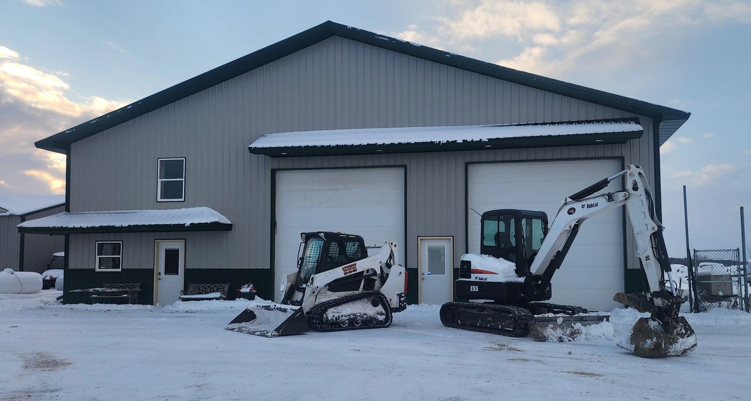Snowy scene: a large gray building with garage doors, a Bobcat skid-steer and excavator sit outside.