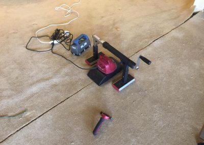 Machine on carpet, powered by an electrical cord, with a pink hand-held device on the floor.