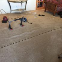 Black line cut through worn beige carpet, with a tool resting on it. Furniture in background.