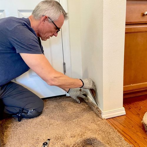 Man kneeling, installing baseboard on beige carpet near doorway and cabinet.