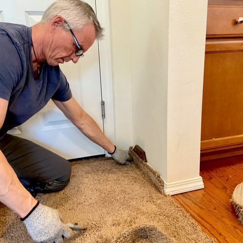 Man kneels, removing carpet from a wall corner near a door. He wears glasses, gloves, and dark clothing.