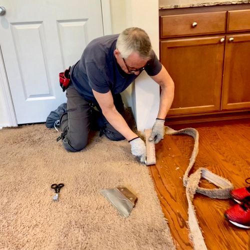 Man kneeling, installing carpet along a wooden floor edge.