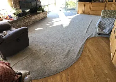 A gray carpet with a curved edge laid over wood flooring in a living room with a recliner and fireplace.