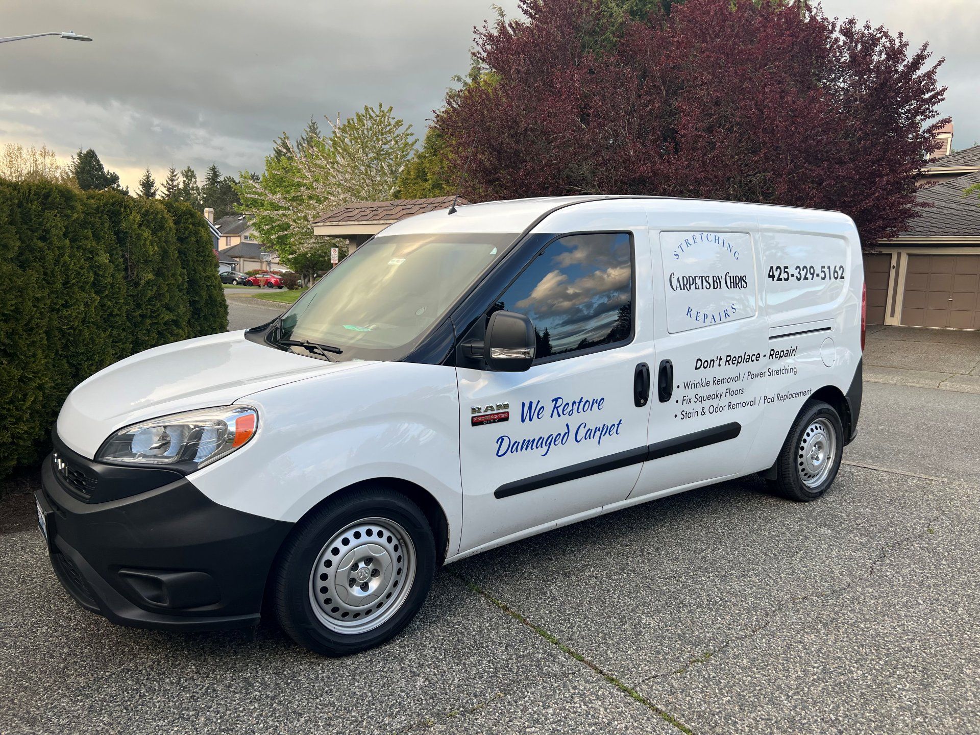 White service van parked on a driveway with company logos and contact info. Trees and a house are in the background.