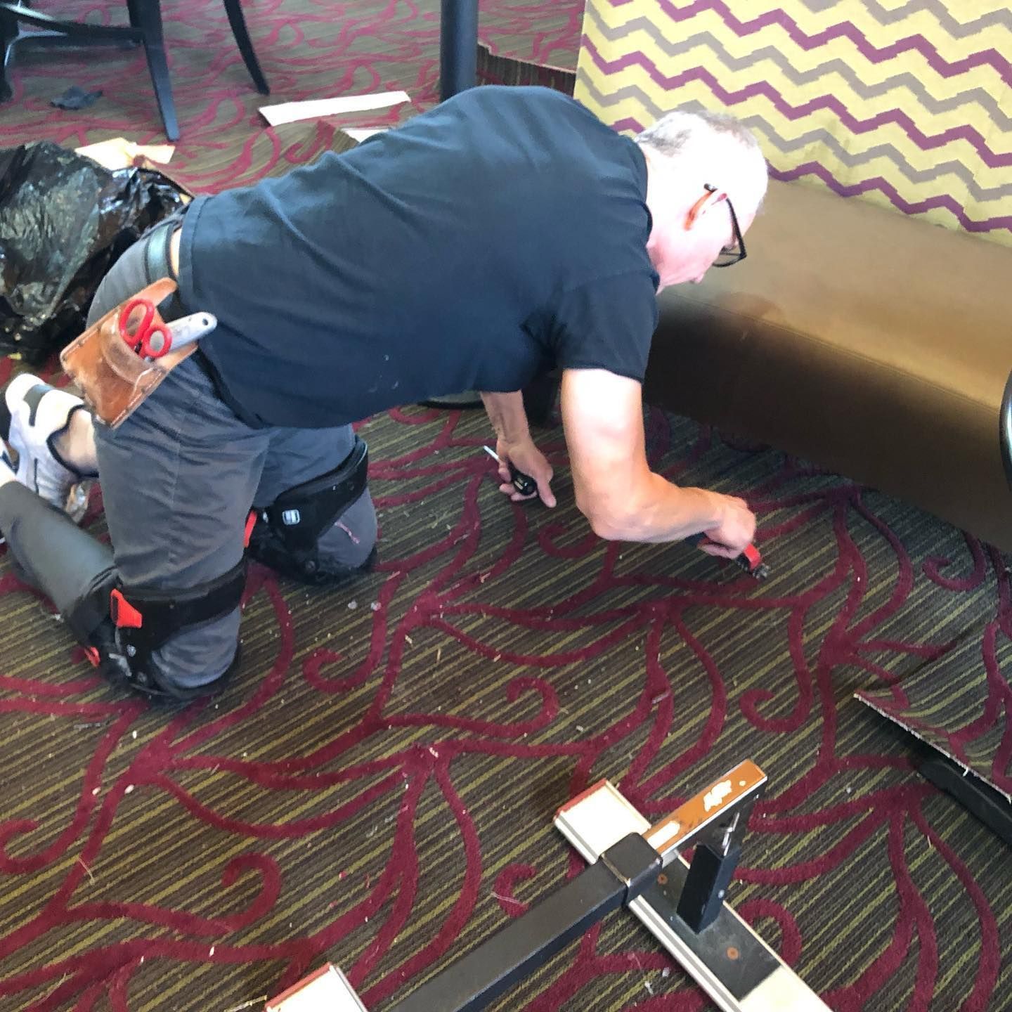 Man on knees cutting carpet with a tool. He wears kneepads, a tool belt, and is in a room with patterned carpet and a brown bench.