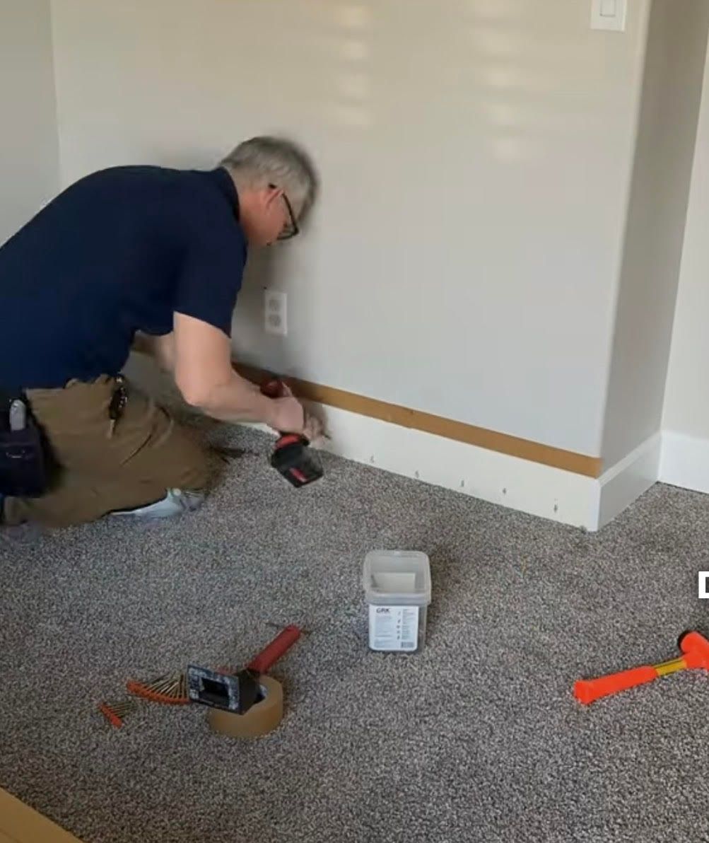Person installing baseboard with a power drill in a room with carpet and beige walls.