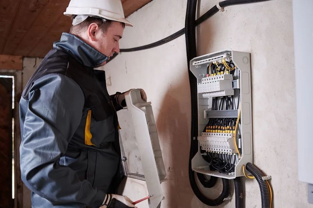Electrician in hard hat working on an electrical panel on a wall.