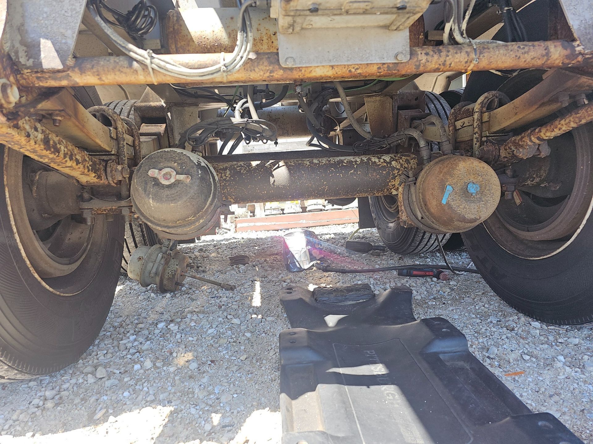 Undercarriage of a rusty truck showing axles, wheels, and brake components on a gravel surface.