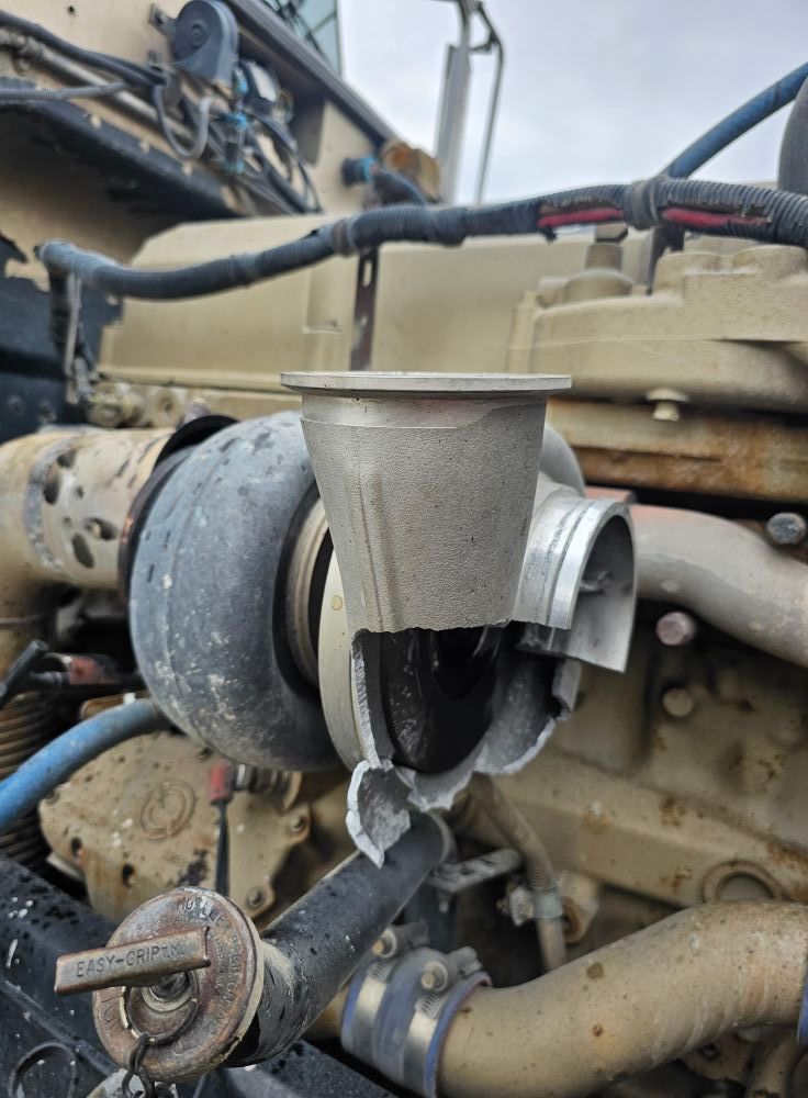 Damaged turbocharger on a tan-colored truck engine. The housing is broken, revealing internal components.