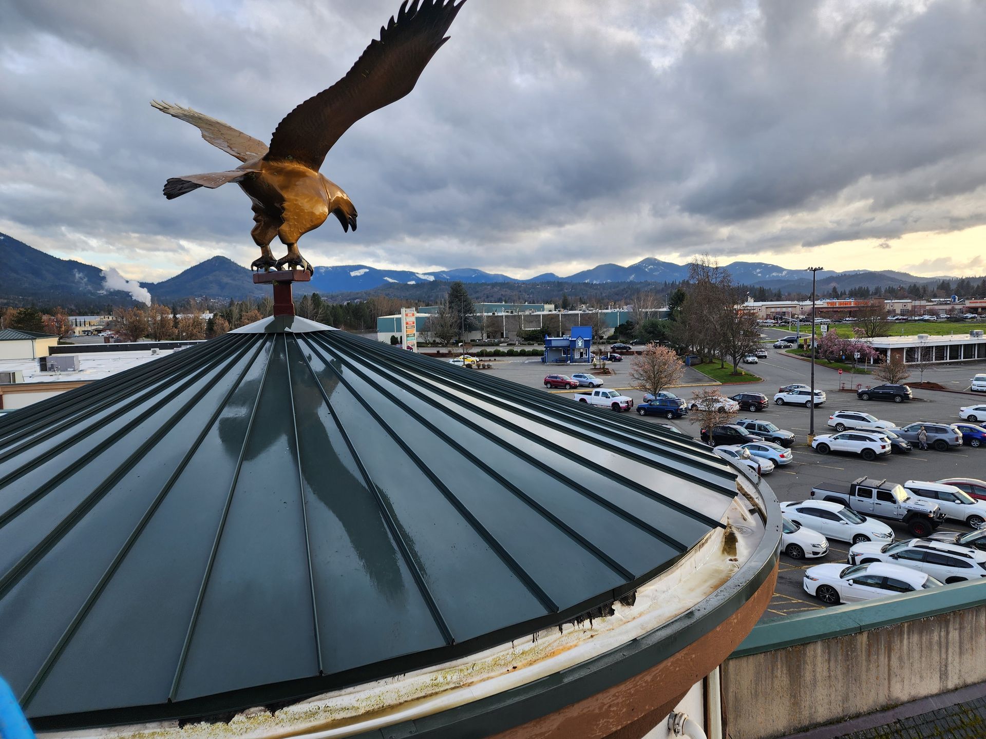 Golden eagle statue atop a green-roofed building, overlooking a parking lot, mountains, and cloudy sky.
