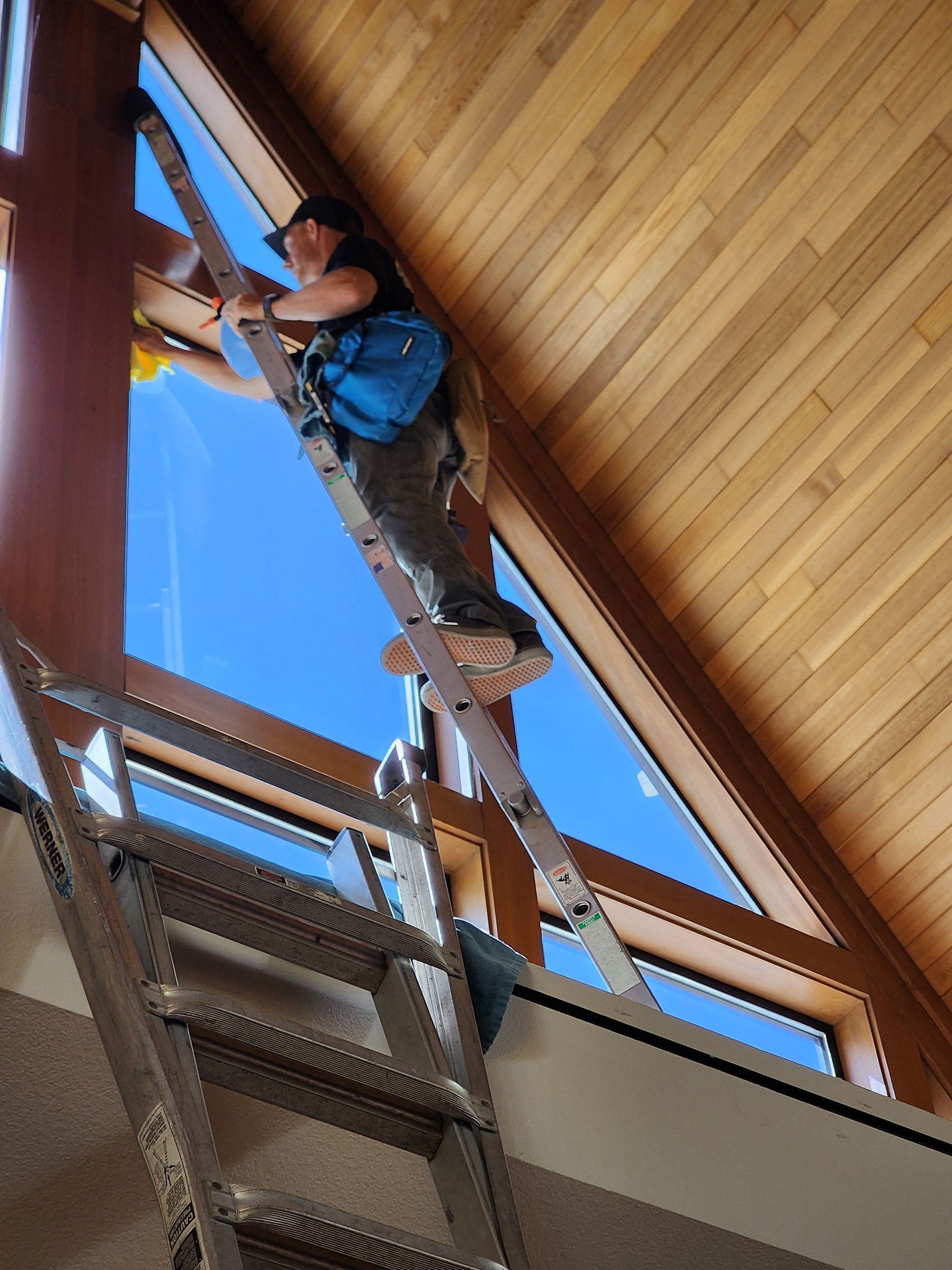 Person on ladder, cleaning a large window with a blue sky visible, inside a wooden building.