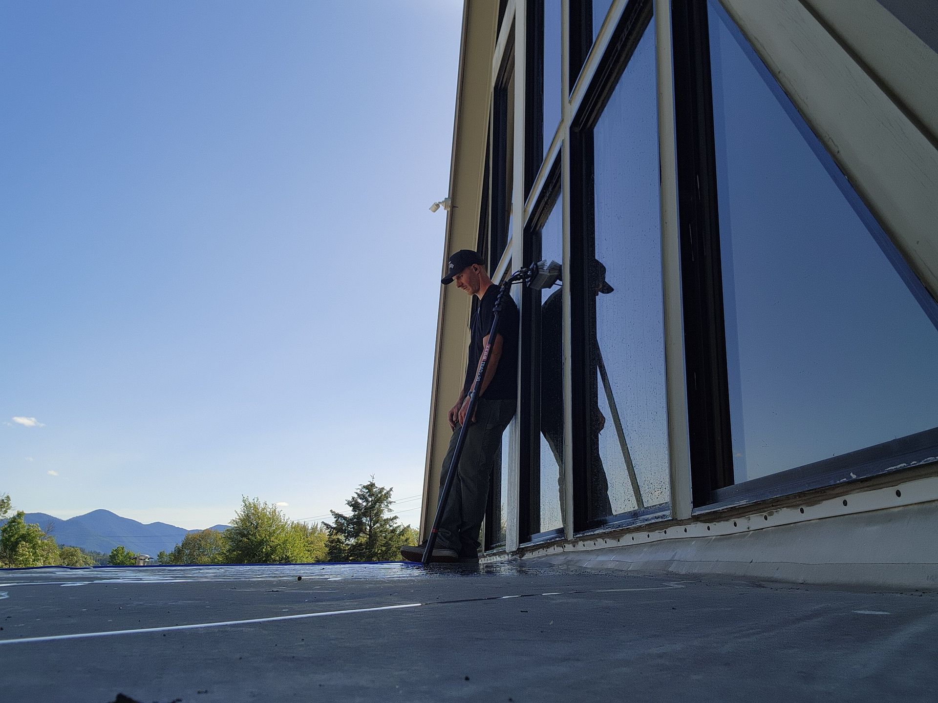 Man cleaning large windows on a building exterior; bright sunlight, clear blue sky; mountain backdrop.