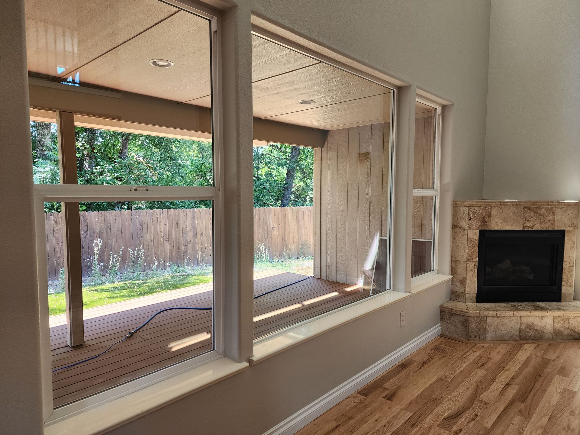 Interior view: large windows looking out onto a deck and trees, beside a stone fireplace and hardwood floor.