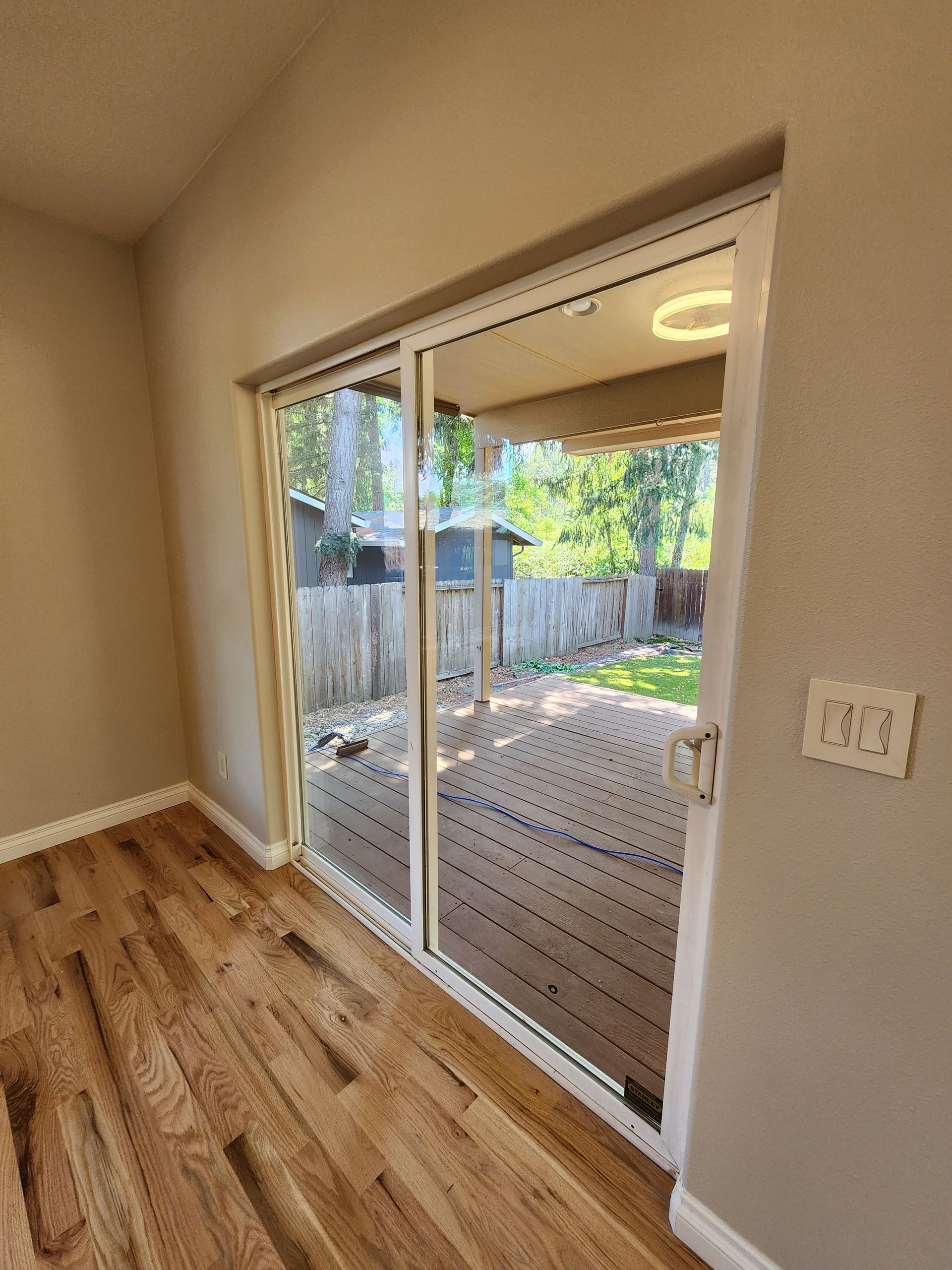 Sliding glass door to a wooden deck, with a backyard view. Sunlight streams in.