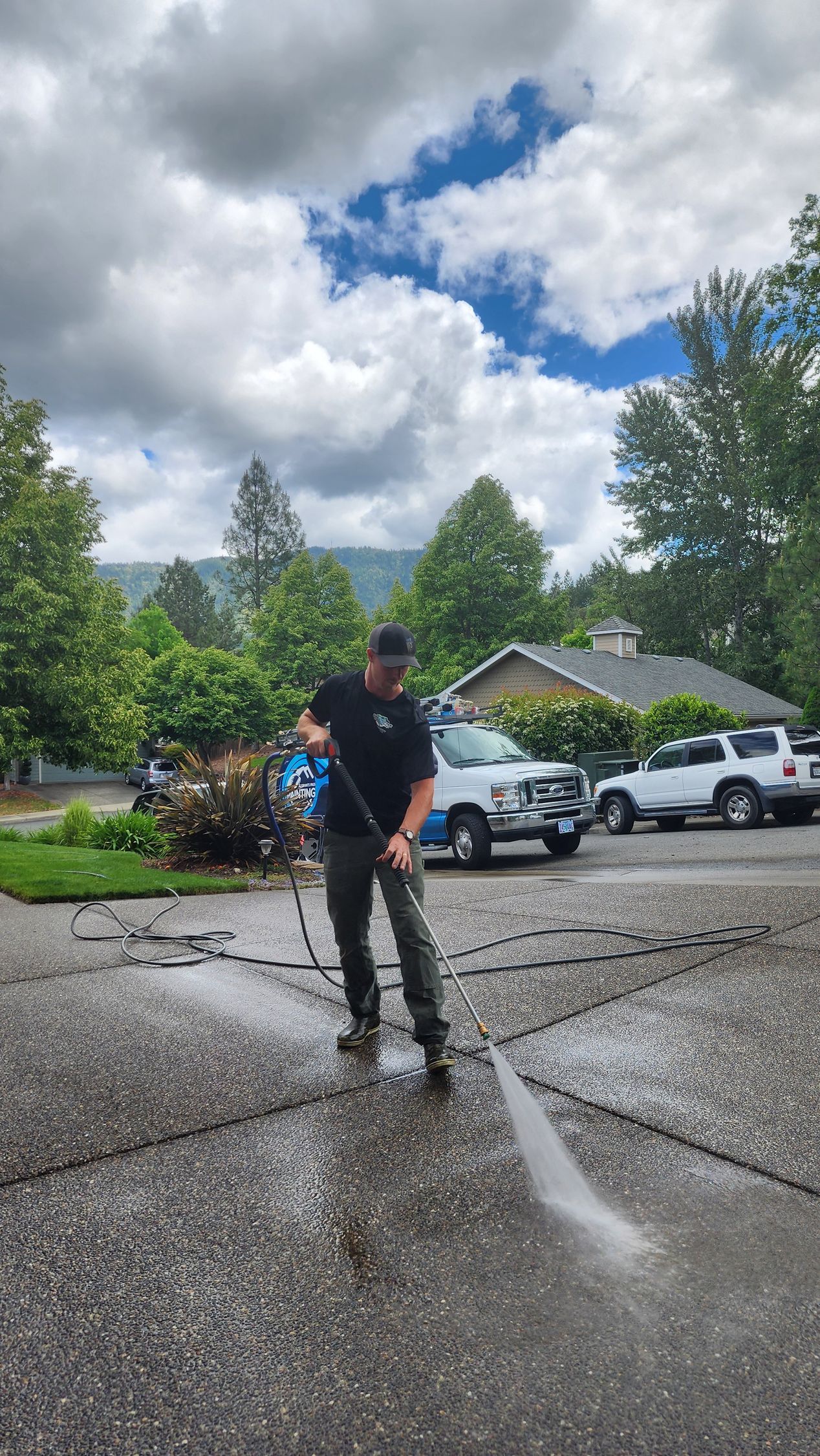 Person pressure washing a driveway on a cloudy day, with vehicles parked nearby and trees in the background.