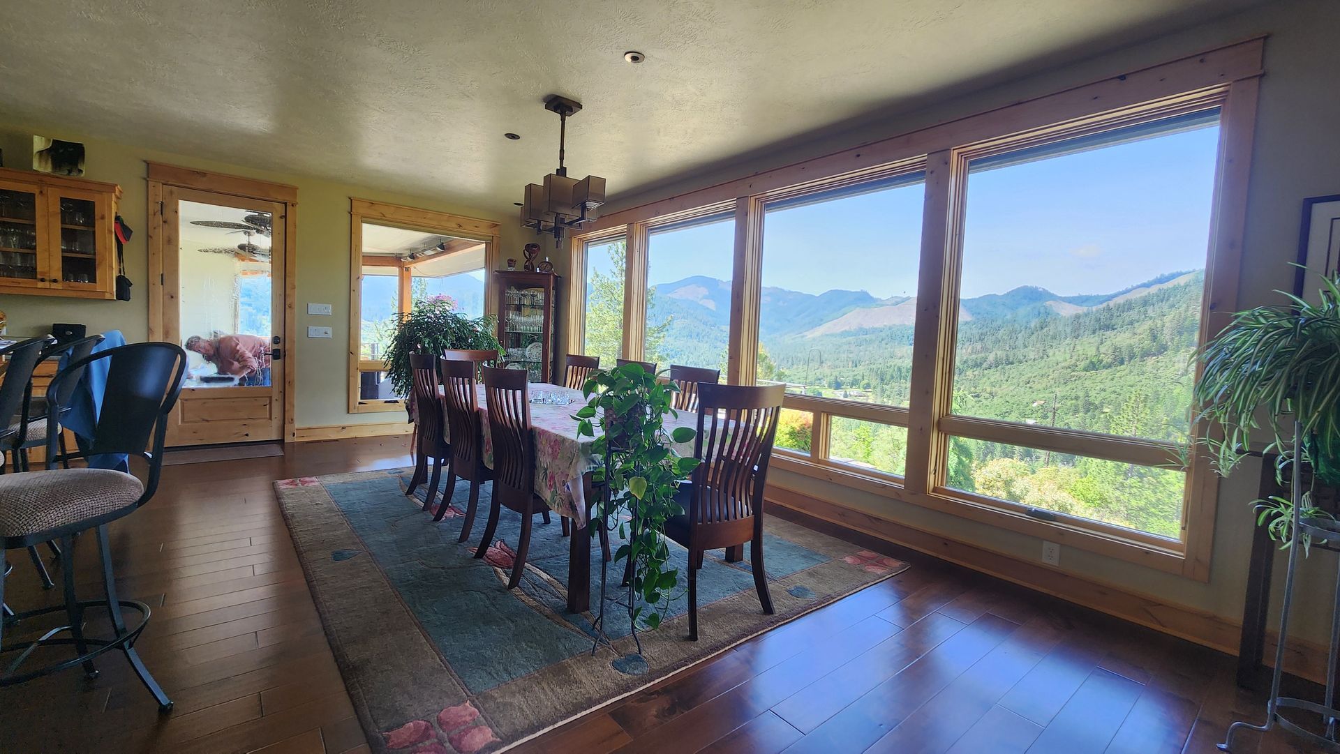 A dining room with a long table, chairs, and large windows with a view of mountains and trees.
