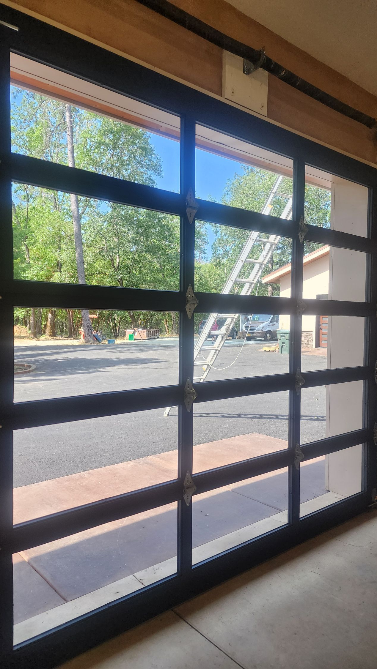Glass garage door with black frame, open to reveal driveway, trees, and a ladder.