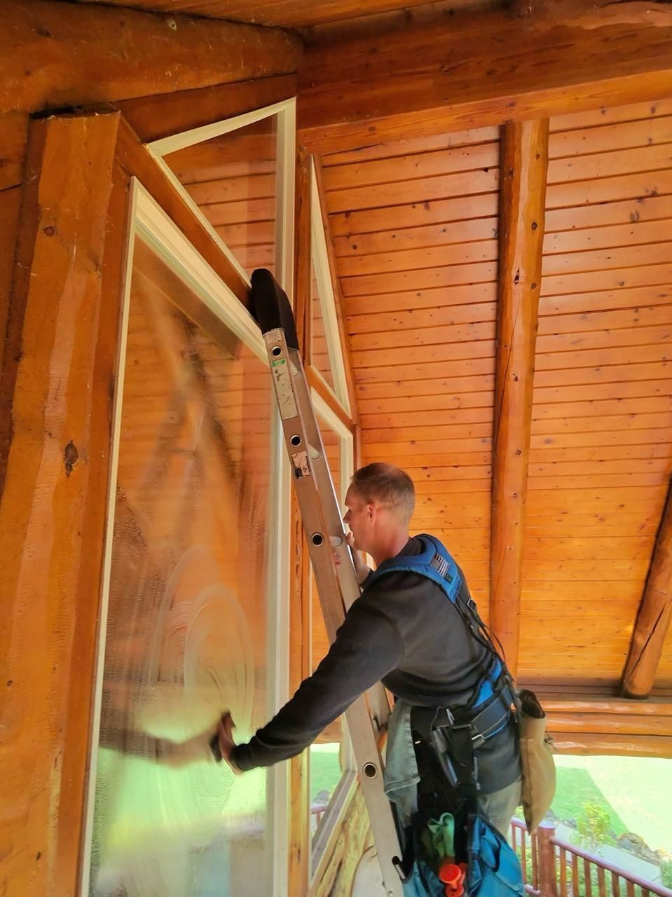 Man on ladder cleaning a large window of a log cabin, wearing safety harness.