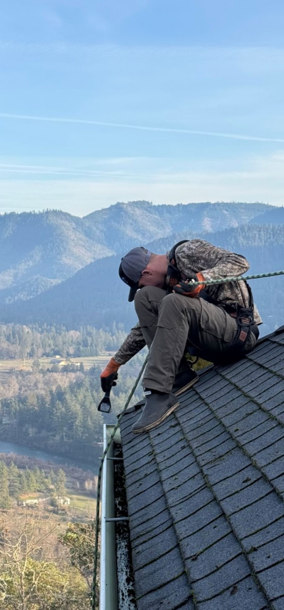 A person cleaning a gutter on a roof with mountains in the background.