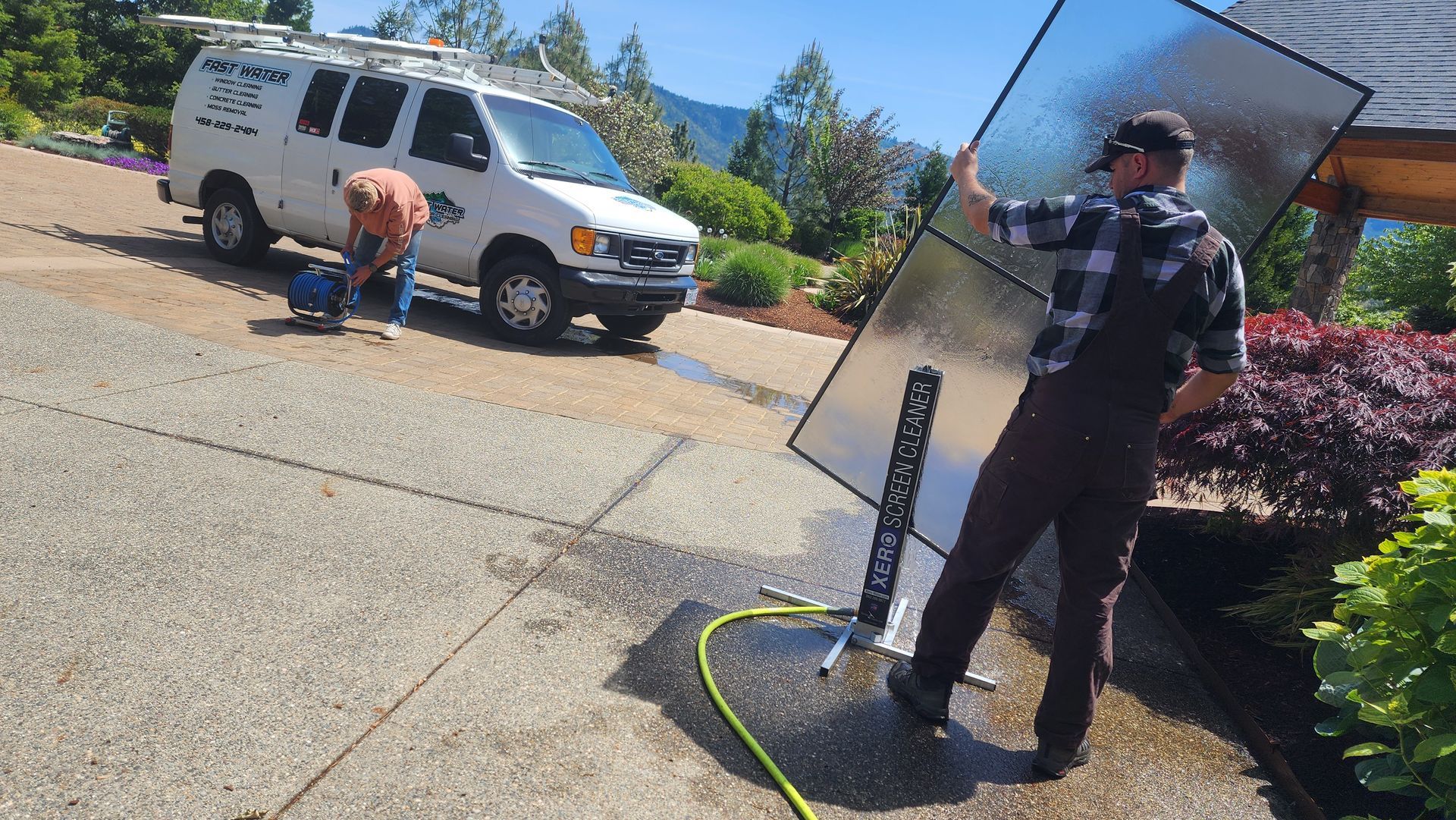 Two men washing a solar panel on a sunny day near a white van and a house with greenery.