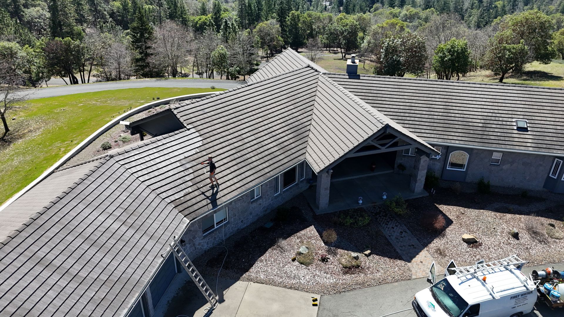 Aerial view of a gray house with a worker on the roof. Trees and a green lawn surround the house.
