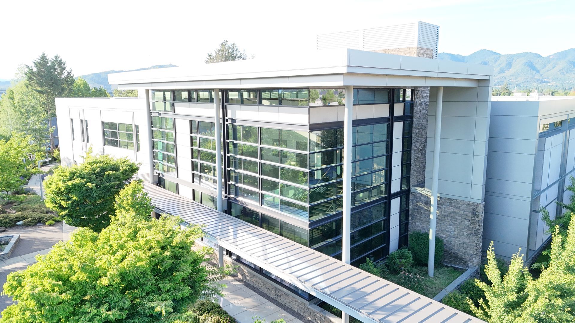 Modern building with glass windows, a walkway, and lush green trees. Mountains in the background.