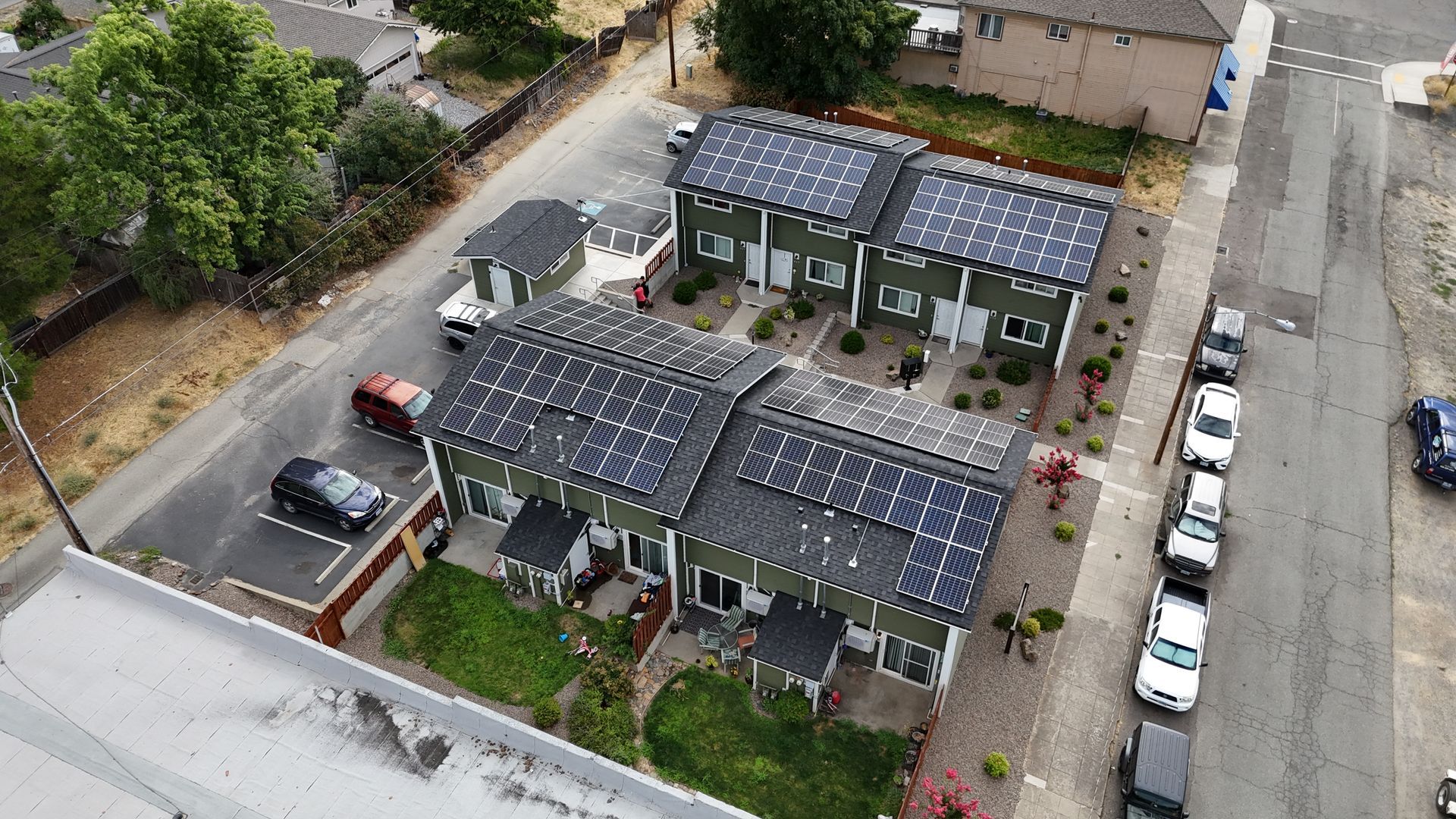 Aerial view of green houses with solar panels, parked cars, and a street.