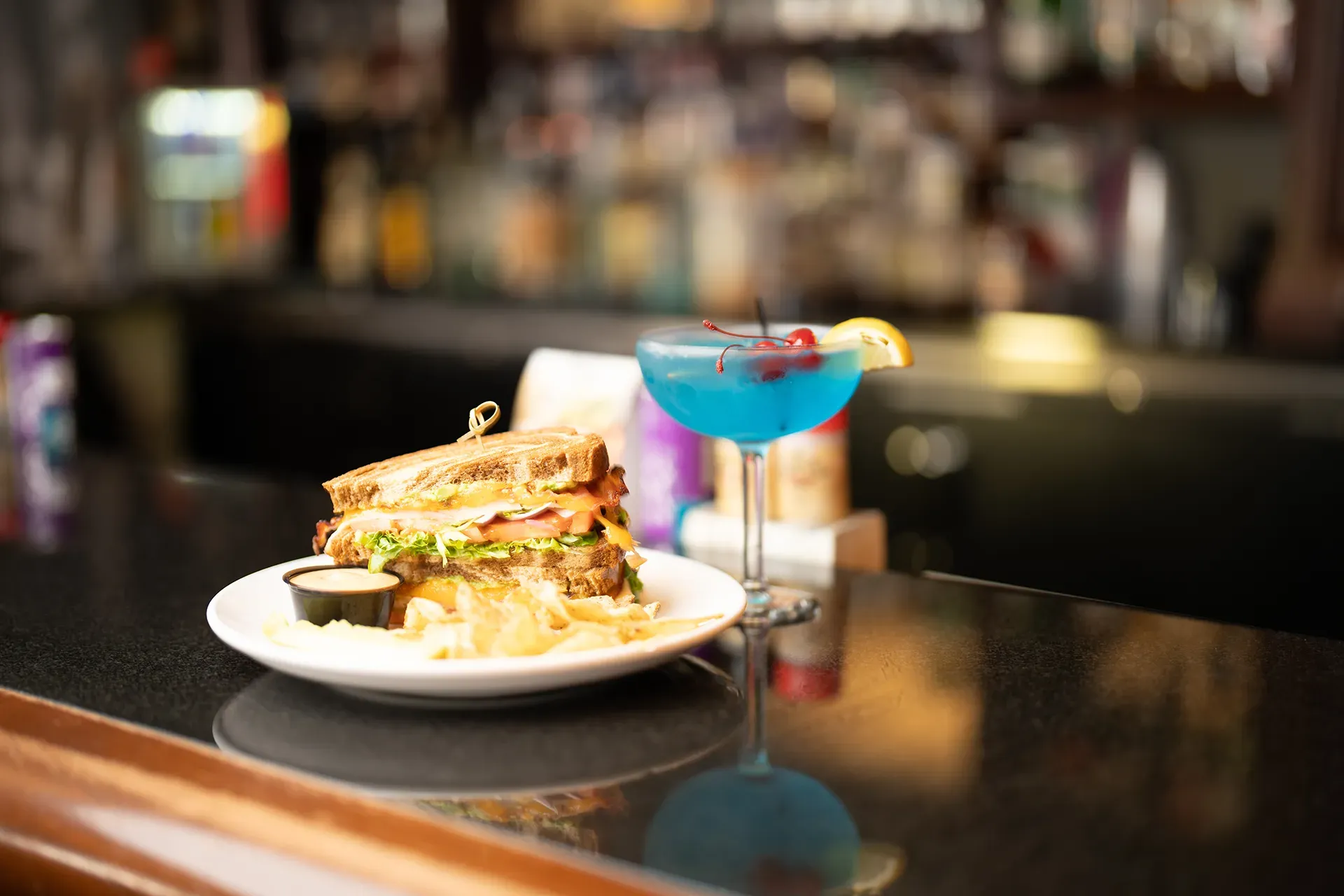 A sandwich and a blue cocktail are on a bar.