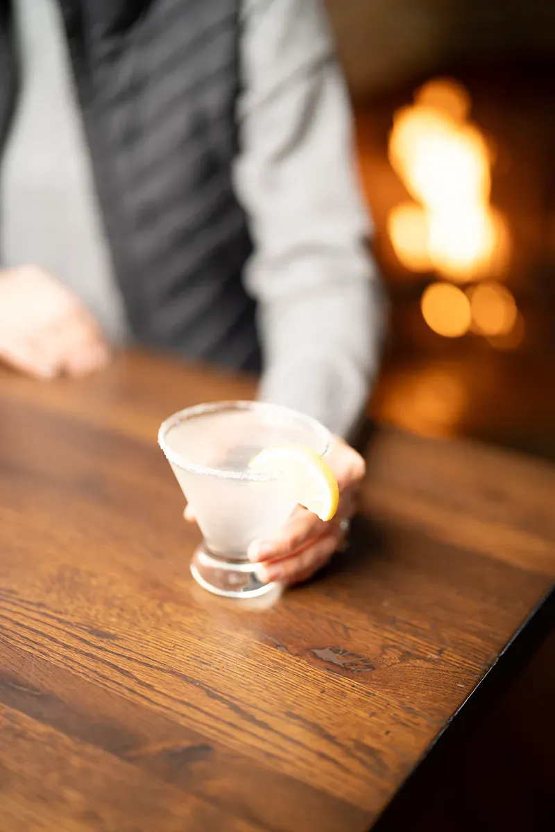 A person is holding a martini with a slice of lemon on a wooden table.