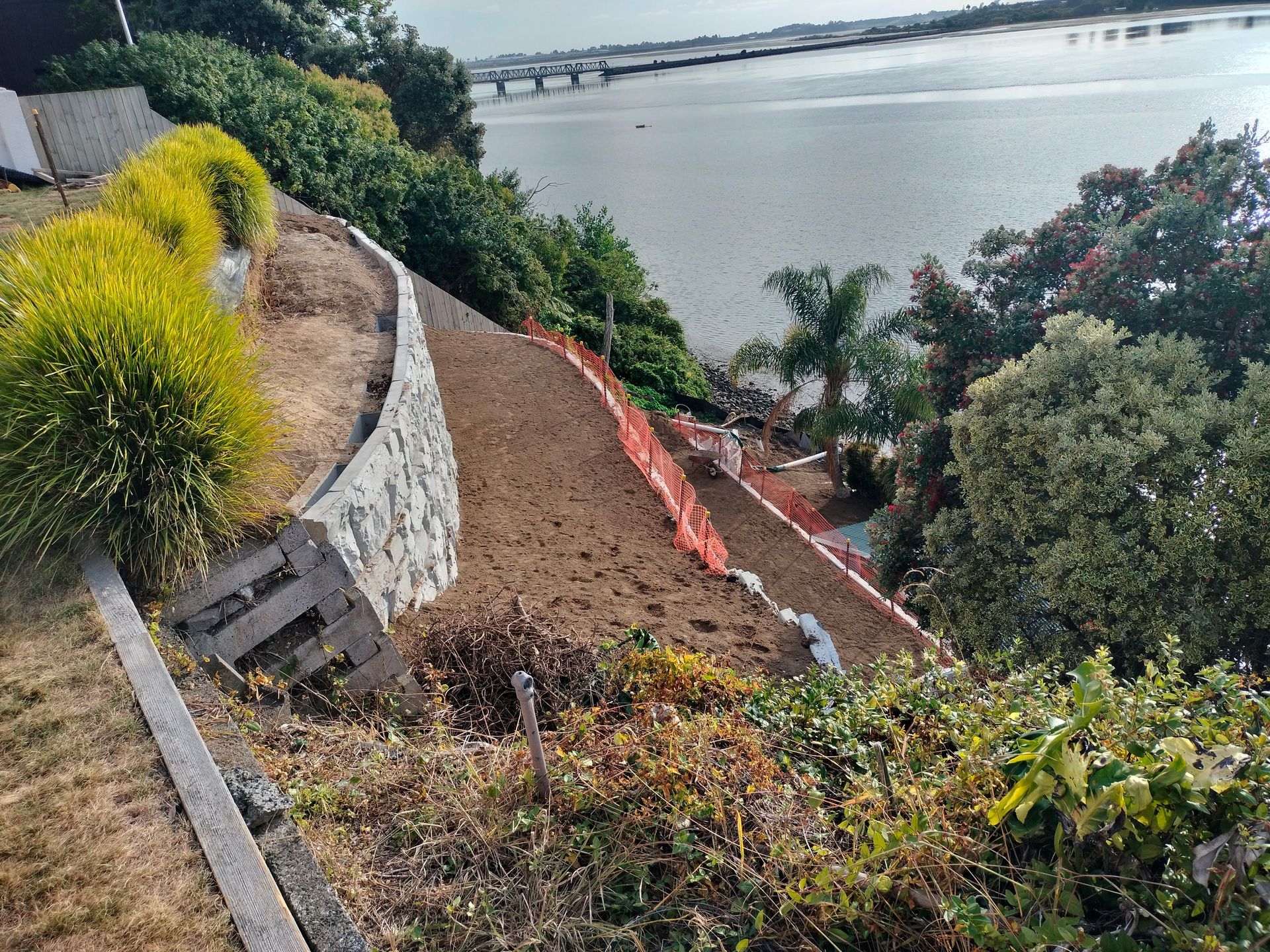 A red wheelbarrow with a shovel stands on a green lawn next to a landscaped garden bed in front of a house.