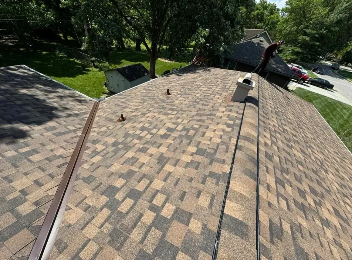 Brown and gray shingle roof on a house, with a chimney and a small shed in the background.