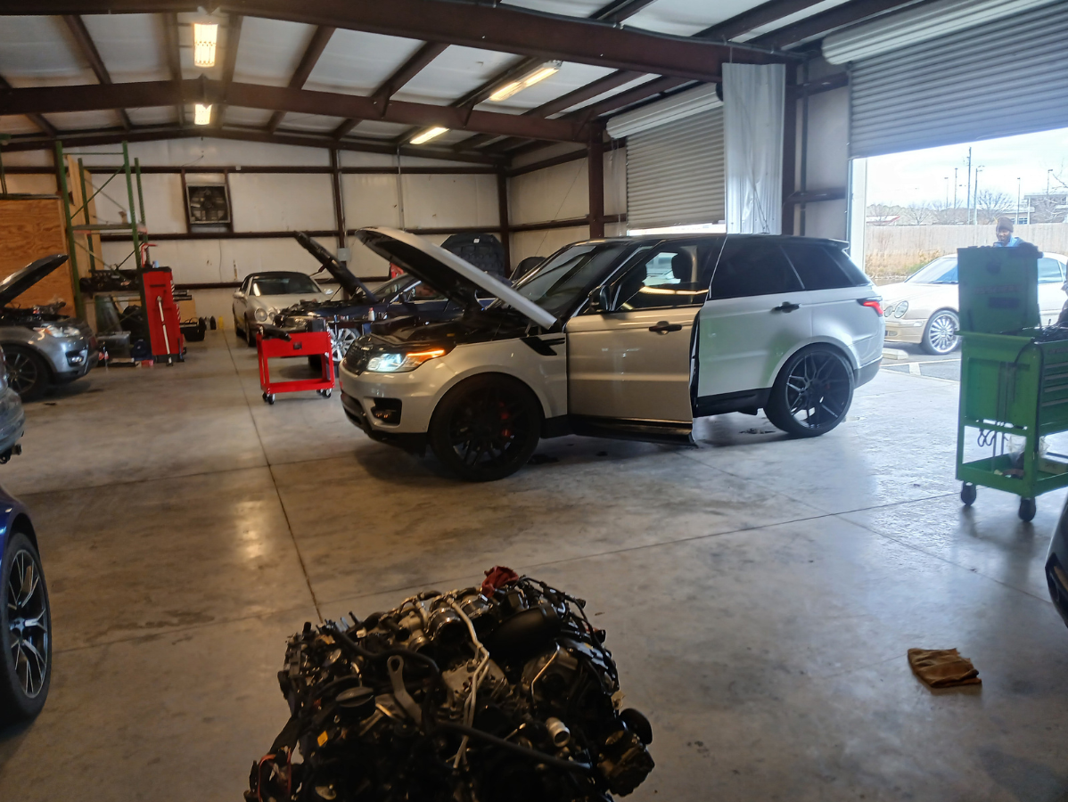 A brightly lit auto repair shop showing a white Range Rover with its hood open and a large engine block in the foreground.