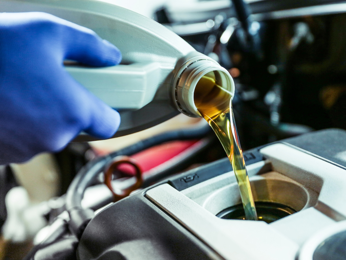 A hand wearing a blue glove pours golden engine oil from a plastic container into a car engine.