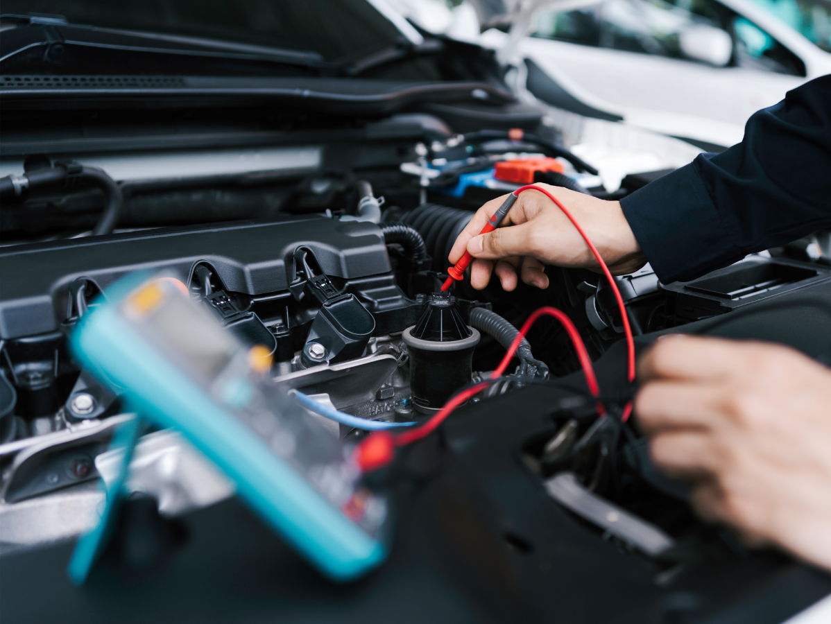 A mechanic uses a digital multimeter to test a component in an open car engine bay.