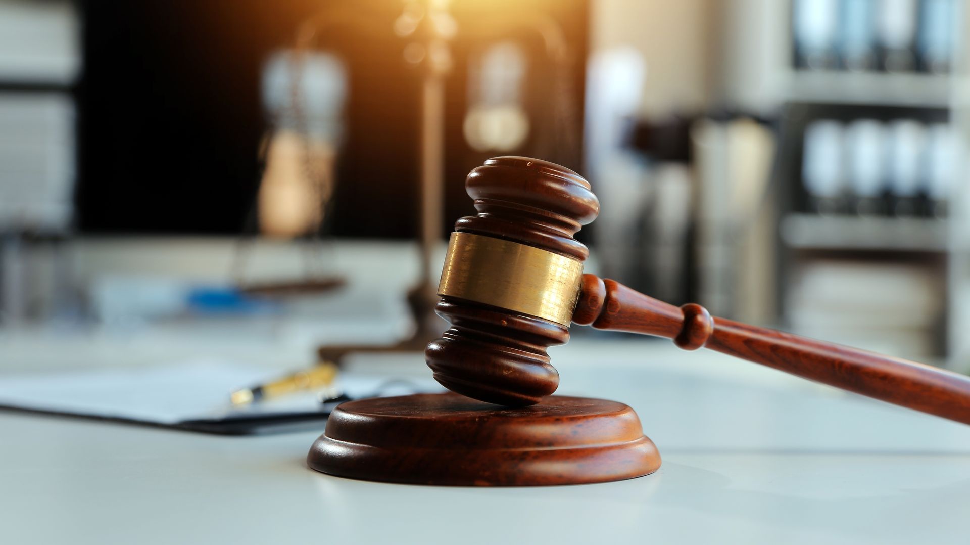 Wooden gavel resting on a sound block on a desk. A blurry backdrop includes a scale and documents, in a law office.