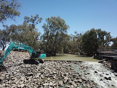 A large excavator is sitting on top of a pile of rocks next to a river.