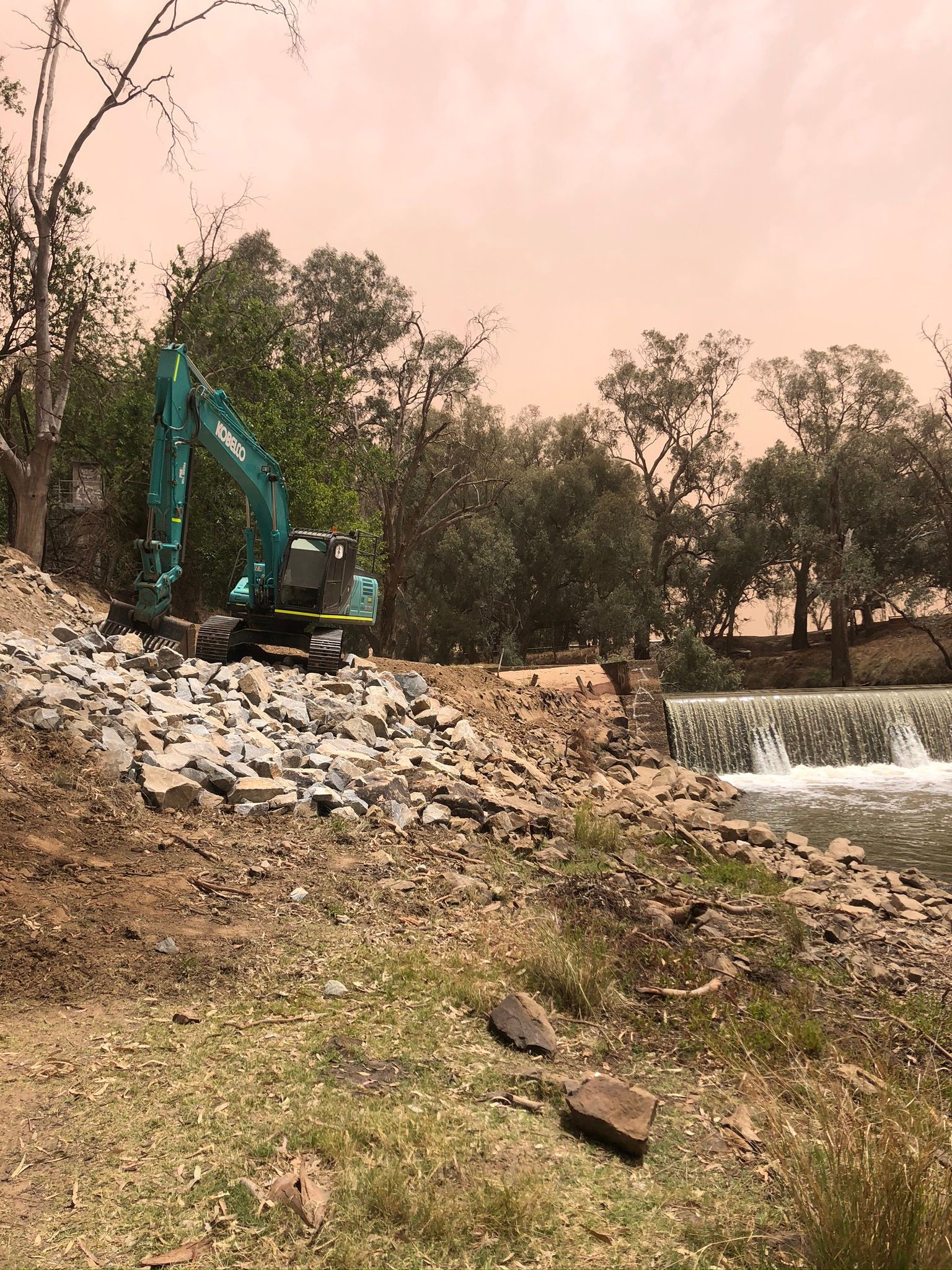 A large excavator is sitting on top of a rocky hill next to a river.