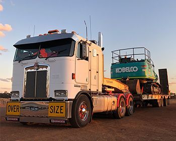A white truck is carrying a green kobelco excavator on a trailer.