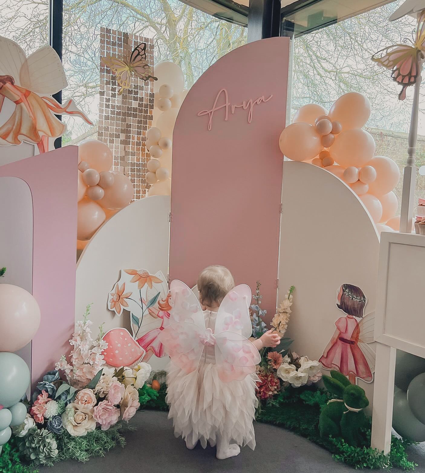 A little girl in a pink dress with wings is standing in front of a wall with balloons and flowers.