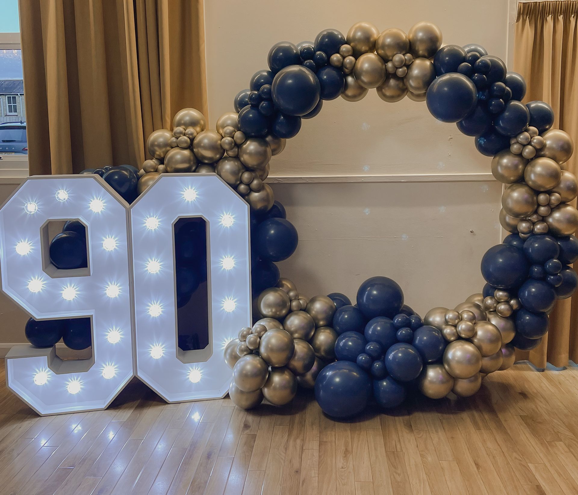 A large number 90 is surrounded by balloons in a room.