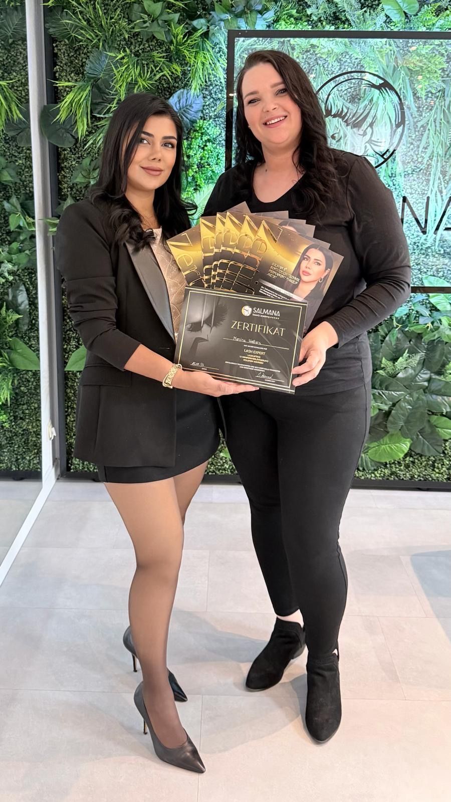 Zwei Frauen in schwarzen Outfits halten ein goldumrandetes Zertifikat in den Händen und stehen im Freien vor grünem Laub.