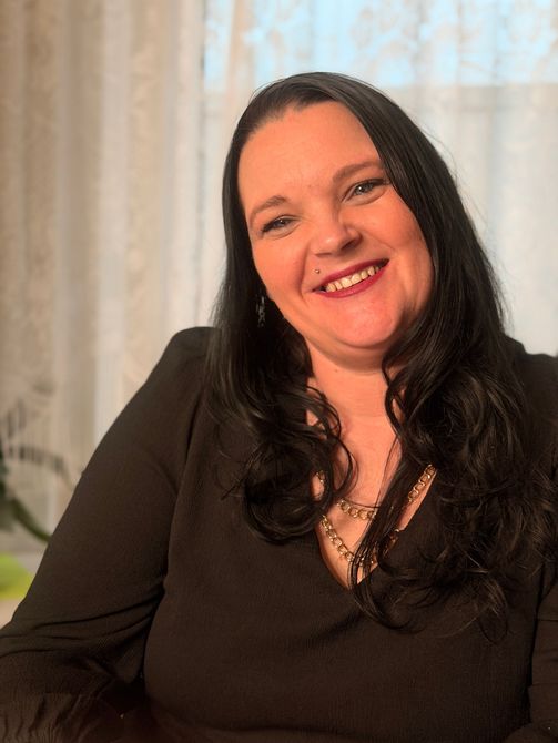 Lächelnde Frau in schwarzem Oberteil sitzt drinnen vor hellen Vorhängen.