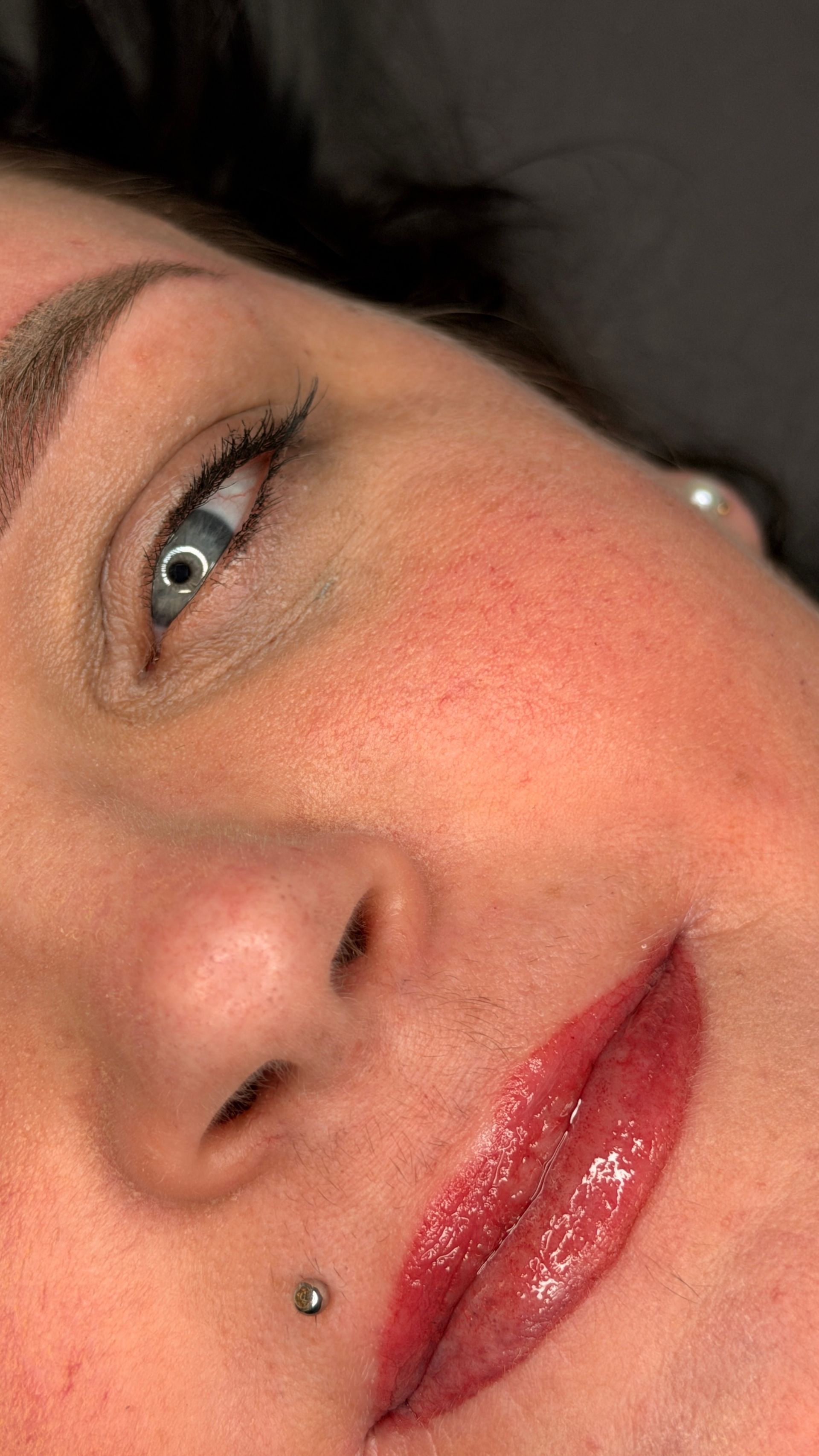 Nahaufnahme eines Frauengesichts mit rotem Lippenstift und kleinem Nasenpiercing, sanft beleuchtet vor dunklem Hintergrund