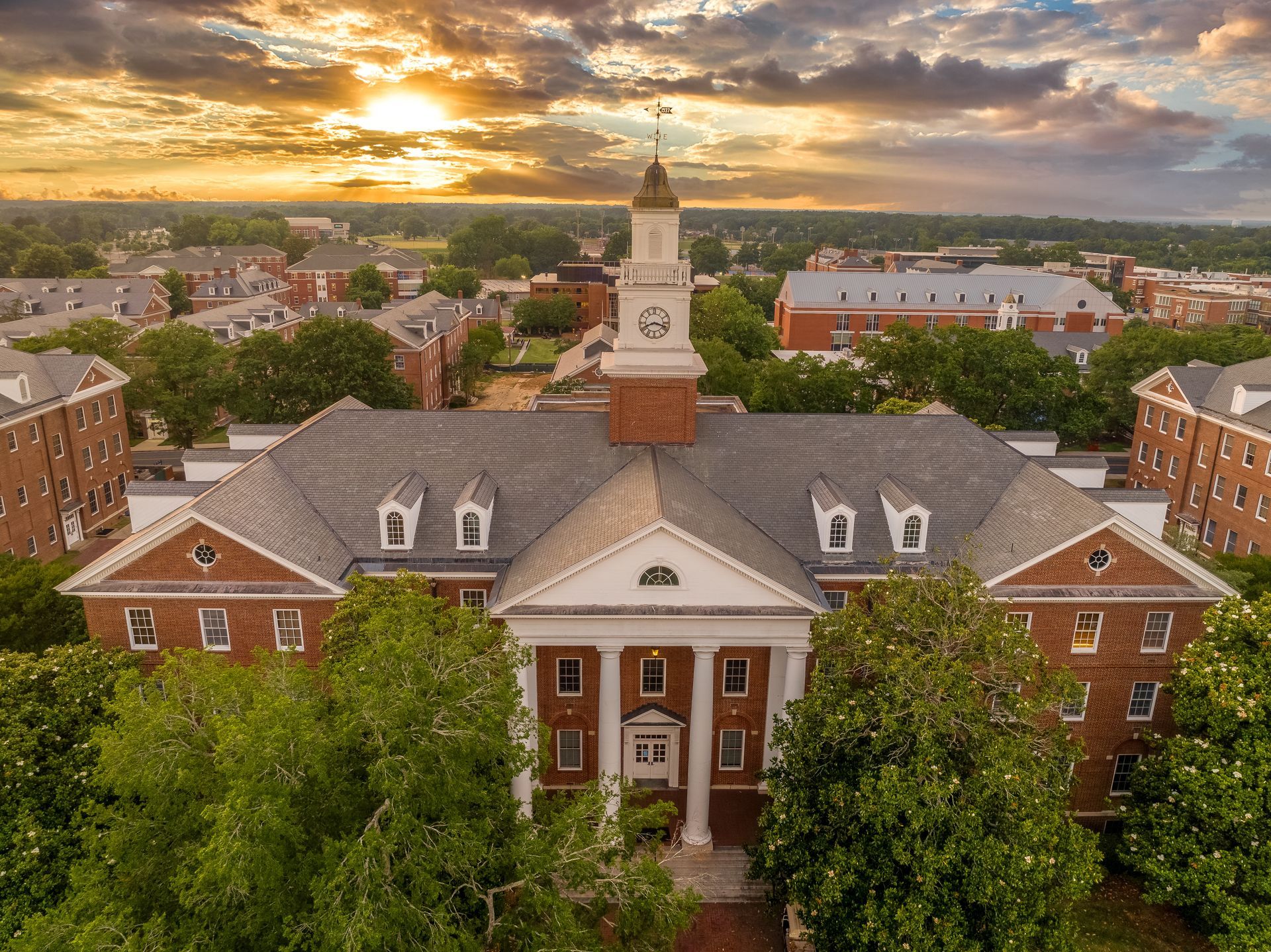 Aerial view of a brick campus building with a central clock tower at sunset, framed by trees.