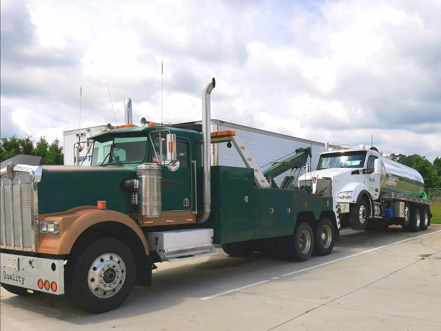 a green tow truck is parked next to a white semi truck .