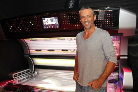 Mann steht neben einem Solarium; lächelnd, graues Hemd, Innenraum.