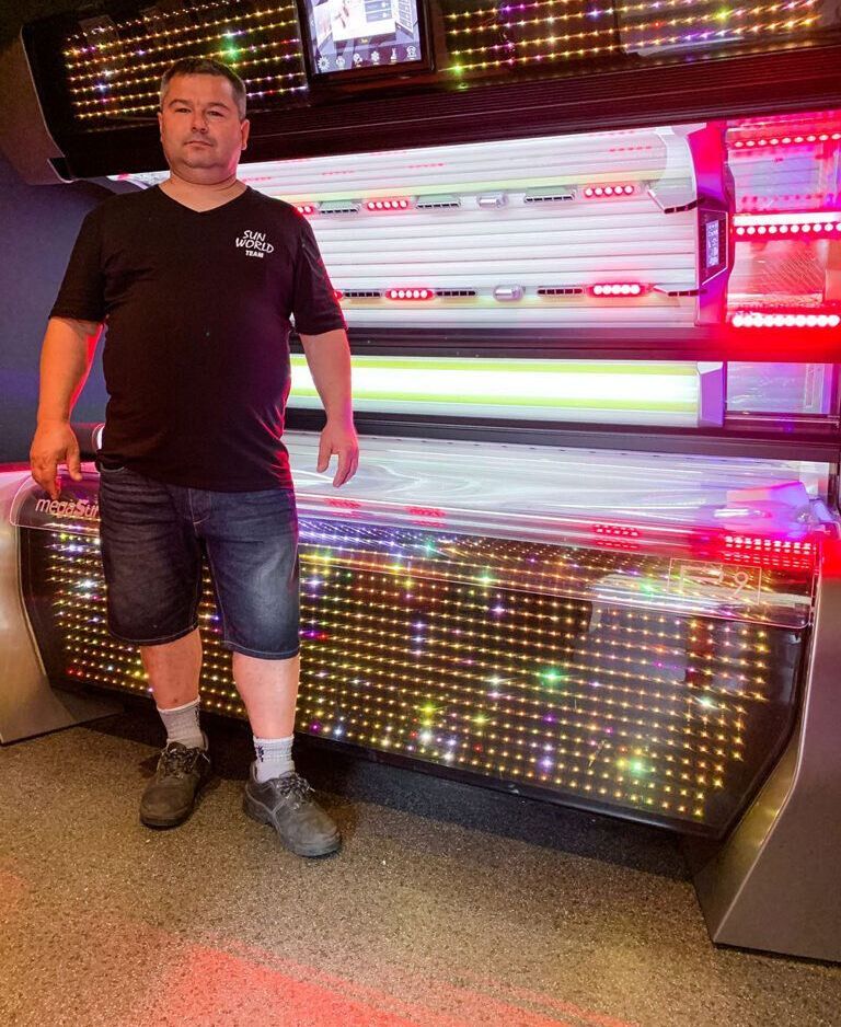 Ein Mann steht neben einer beleuchteten Sonnenbank. Er trägt ein schwarzes Hemd, Jeansshorts und graue Turnschuhe.