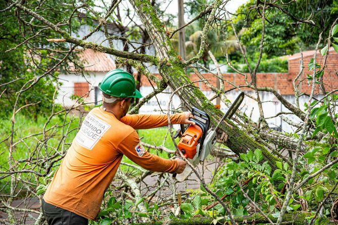 Worker in orange shirt and green helmet cutting branches with a chainsaw near a brick wall